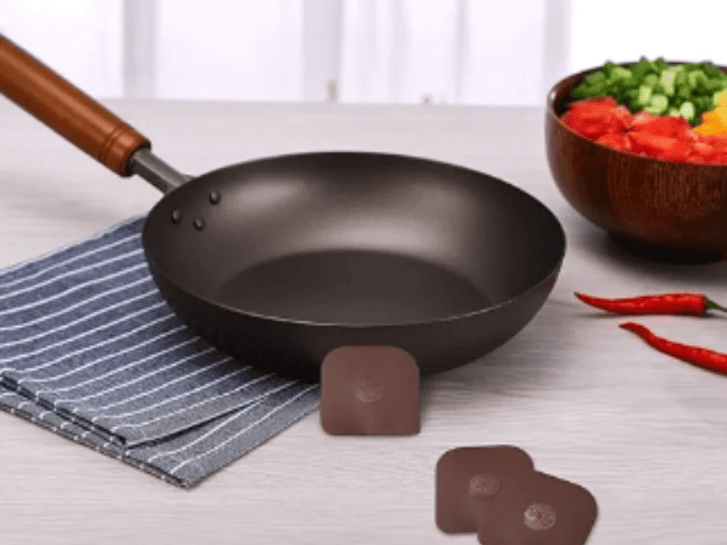 Non-stick frying pan with a wooden handle placed on a striped cloth beside fresh chopped vegetables and red chilies.